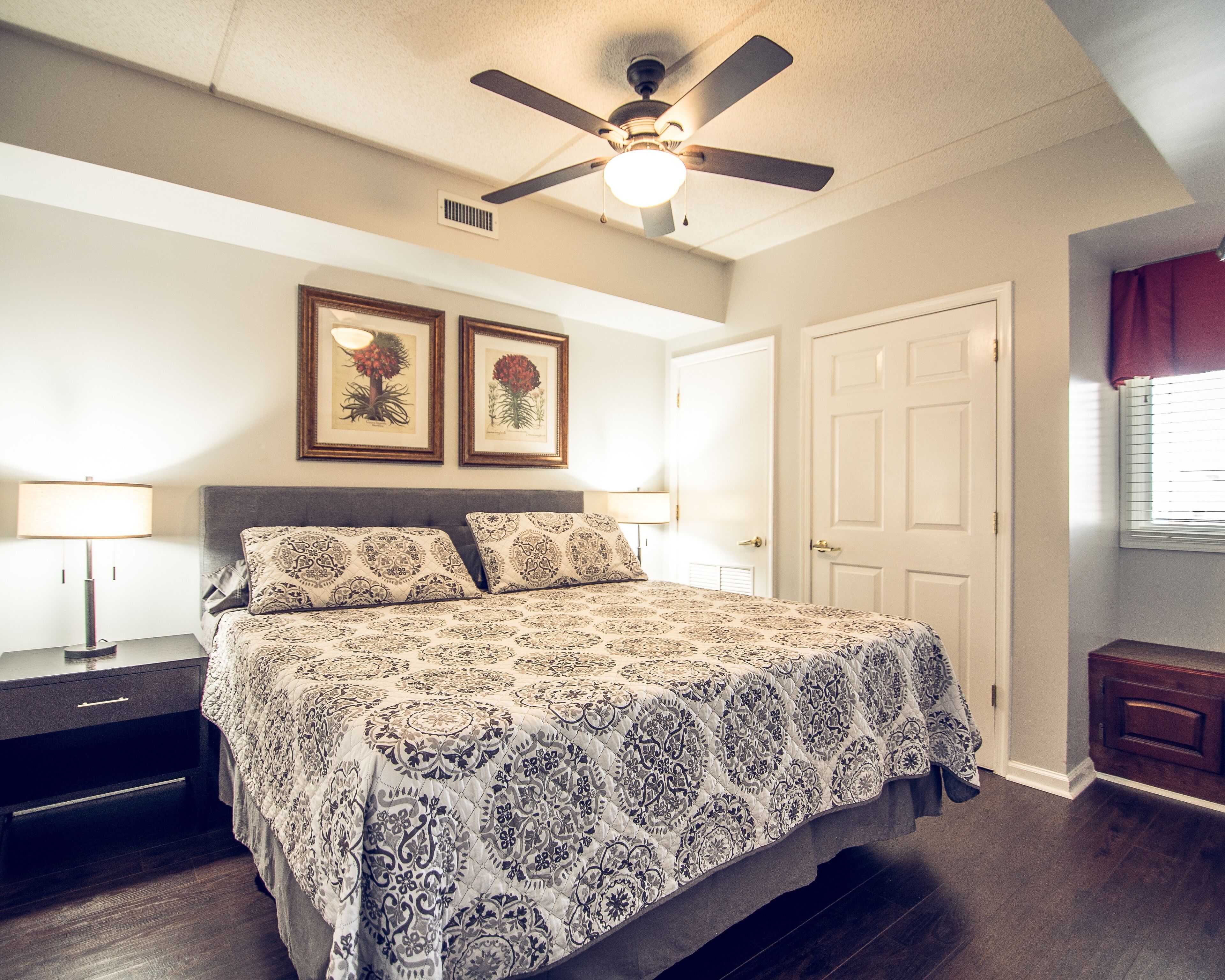A bedroom with a bed, a ceiling fan, a nightstand, a lamp, framed artwork, and a door.