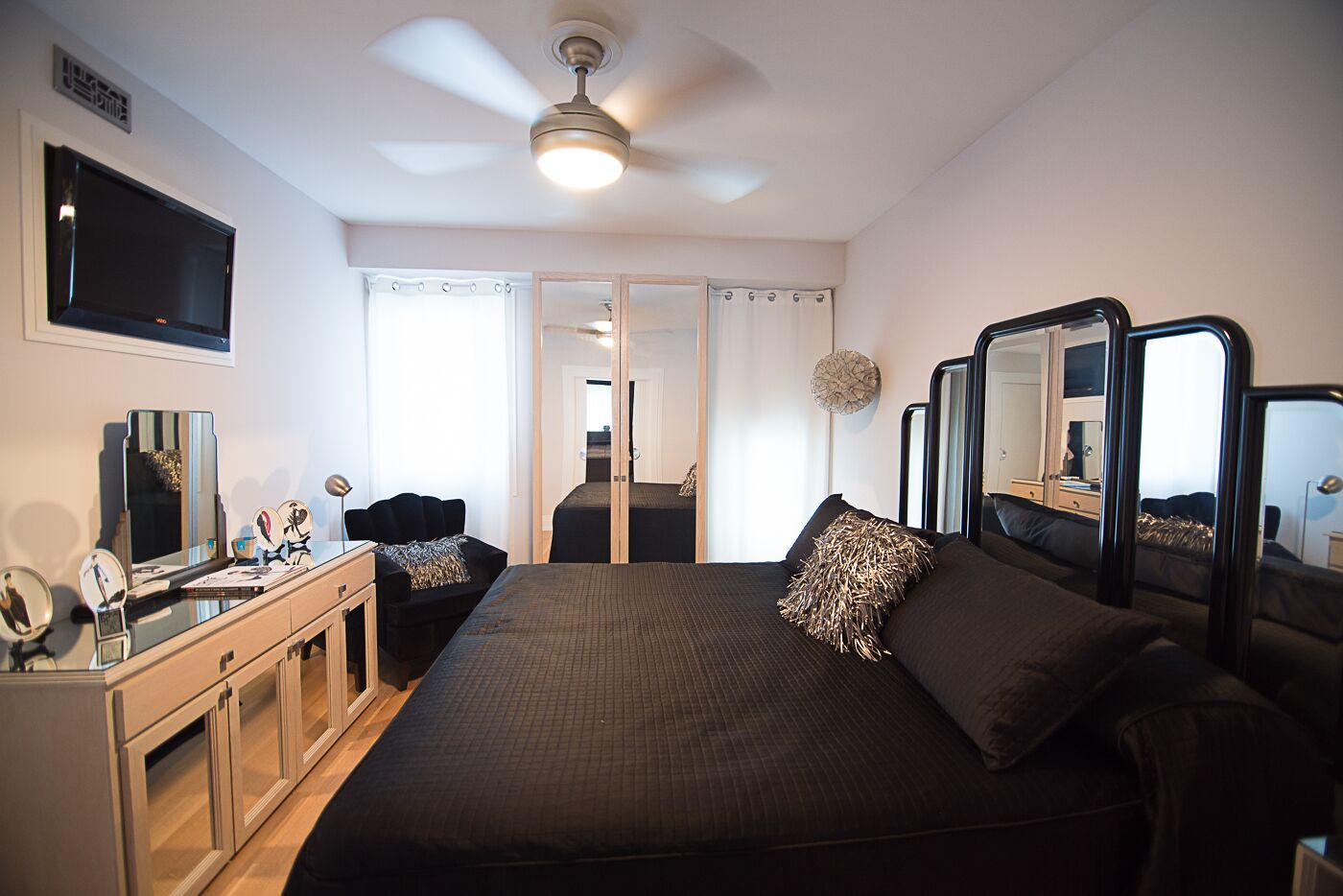 A hotel room with a large bed, bedside tables, a desk with a mirror, a ceiling fan, and a TV mounted on the wall.