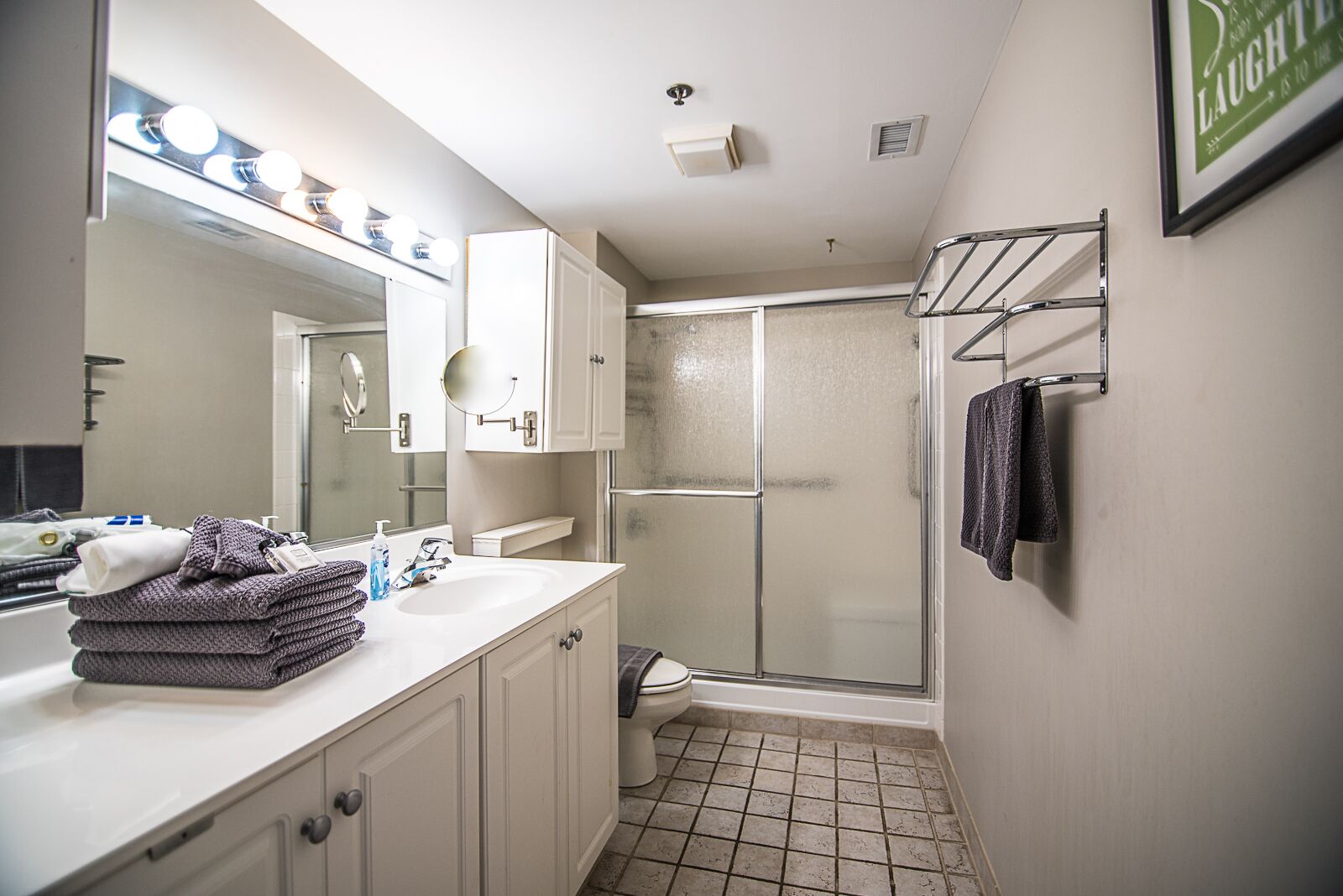 Une salle de bain avec un lavabo blanc, un miroir et une douche. Des serviettes sont pliées sur le comptoir et une autre est suspendue à un porte-serviettes.