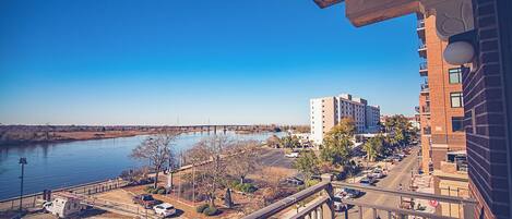 A view from a balcony overlooking a river, a parking lot, and a multi-story building with a clear blue sky.