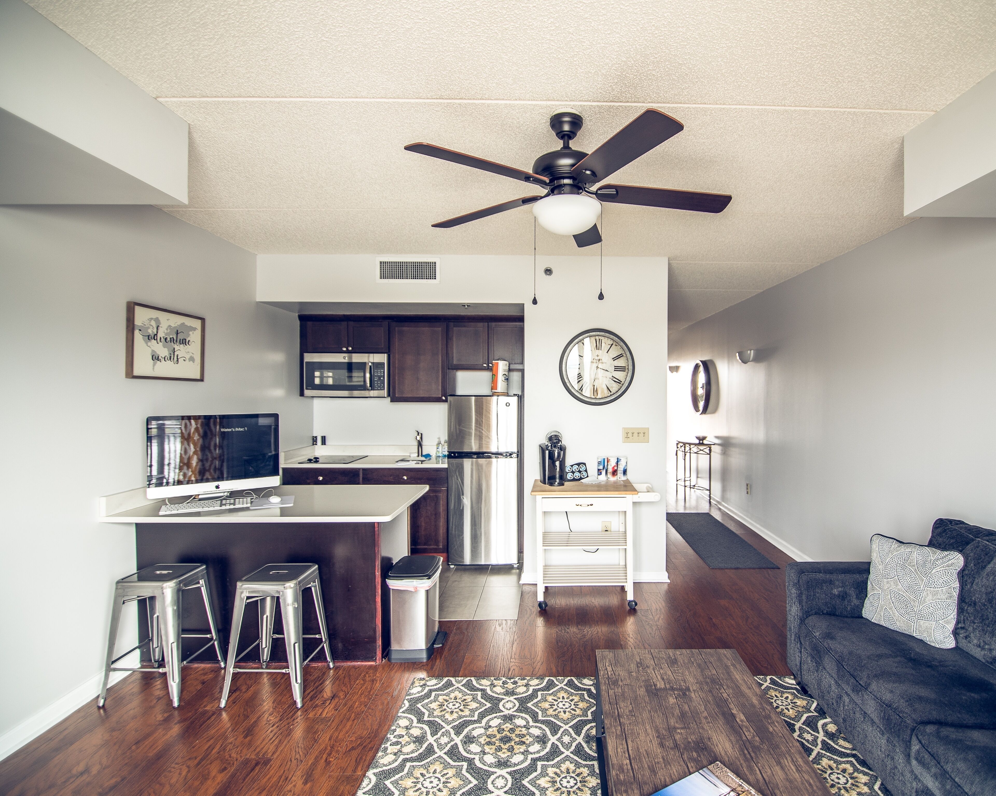 A modern kitchen with dark wooden cabinets, a stainless steel refrigerator, and a microwave. There is a dining area with a white table and two metal stools. A ceiling fan is visible above the kitchen.