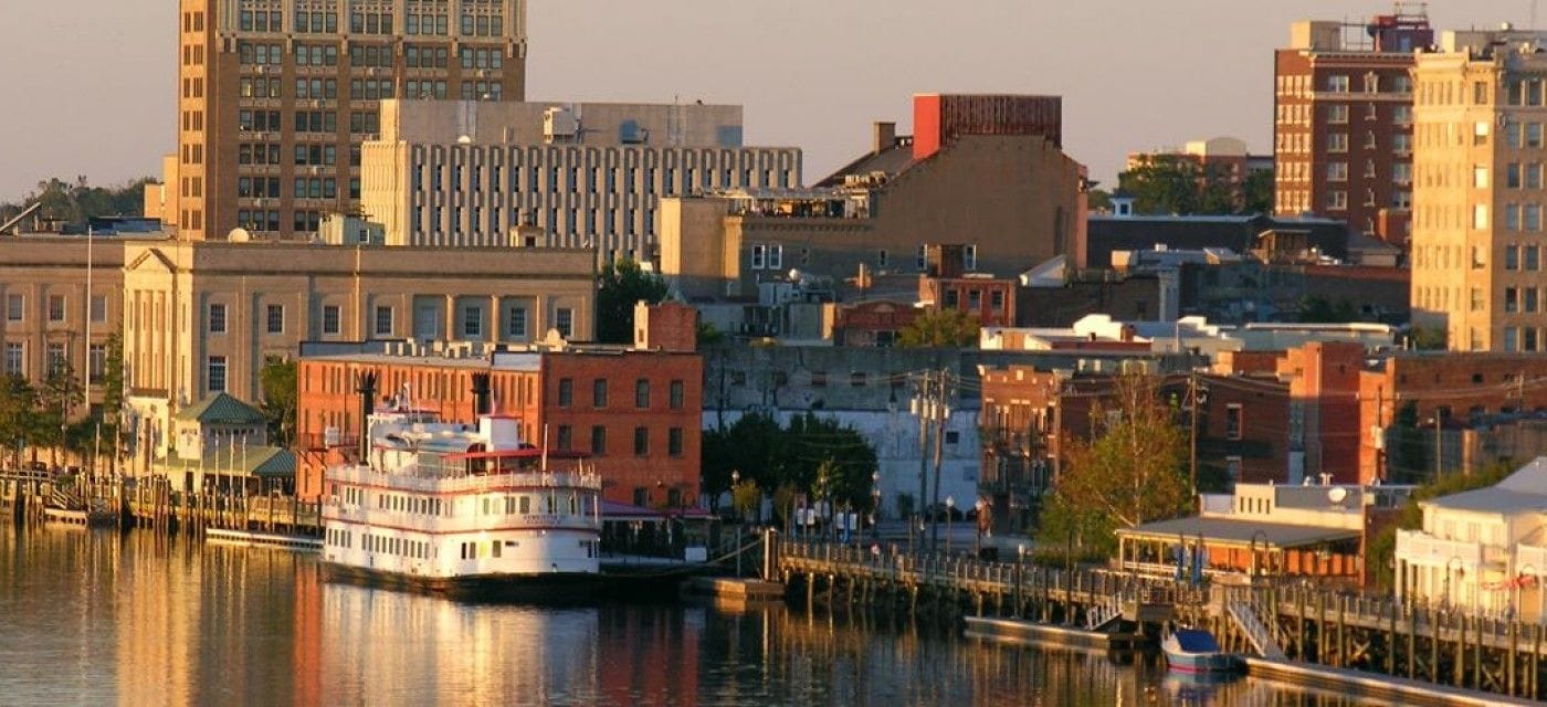A waterfront cityscape with a prominent white riverboat docked at a pier, surrounded by various buildings including a large brick structure and a modern high-rise.