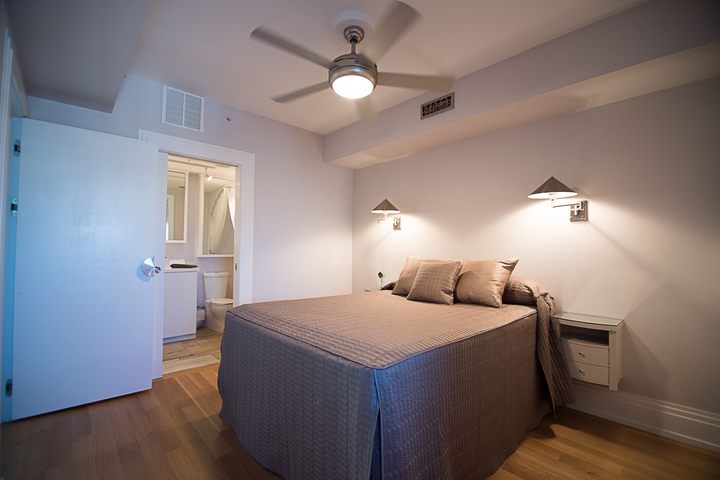 Une chambre à coucher comprenant un lit, des lampes de chevet, un ventilateur de plafond et une porte donnant sur une salle de bain.