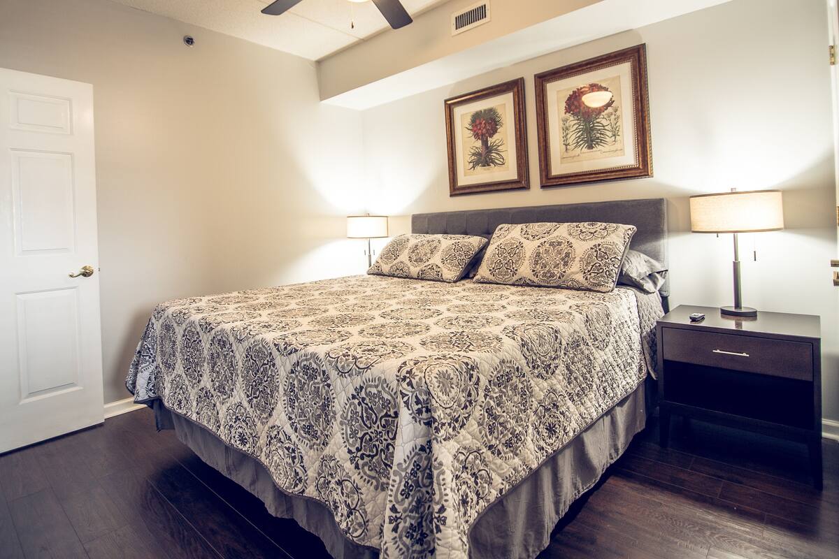 A bedroom with a bed, two bedside lamps, a ceiling fan, and two framed botanical prints on the wall.
