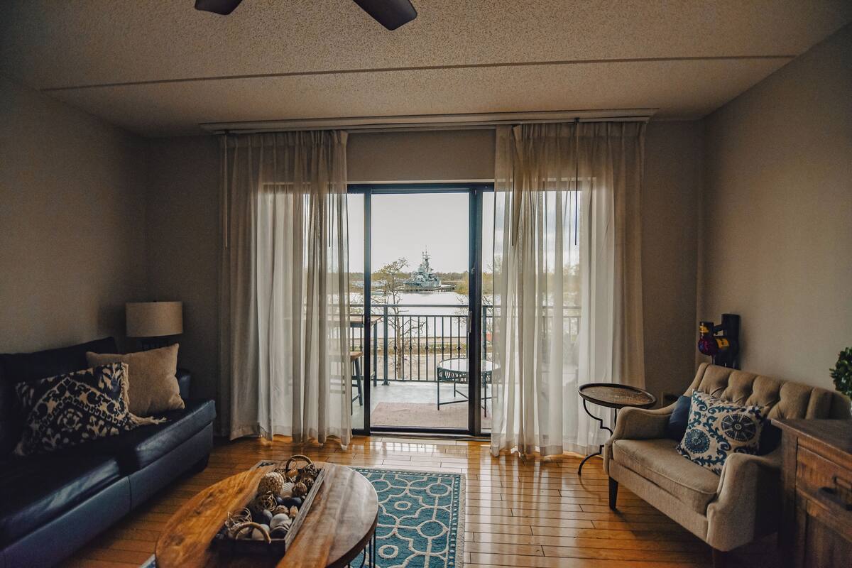 A living room with a sofa, a coffee table, and a chair. There is a large window with curtains, and a balcony is visible through the window.
