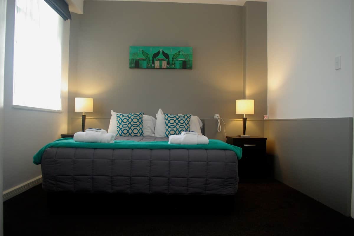 A neatly made bed with a dark quilt and patterned pillows, two bedside lamps, and a wall-mounted artwork above the bed.