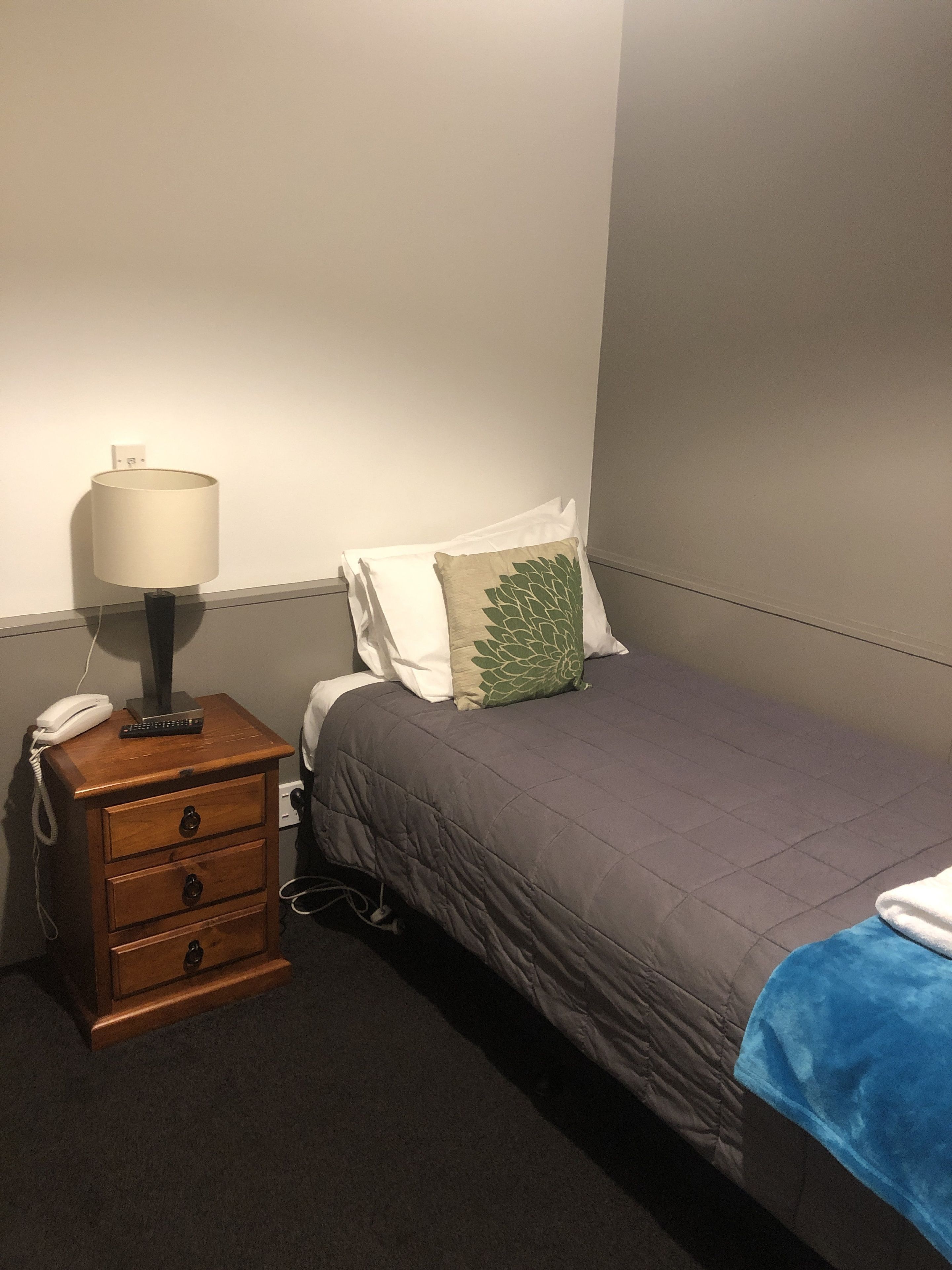 Una cama individual con una manta gris, una mesita de noche de madera con una lámpara, un teléfono y un control remoto.