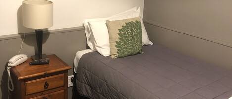 Una cama individual con una manta gris, una mesita de noche de madera con una lámpara, un teléfono y un control remoto.
