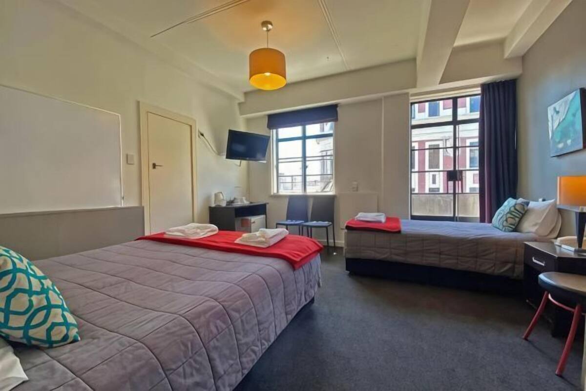 A hotel room with two beds, a desk, a TV, and a window with a view of buildings.