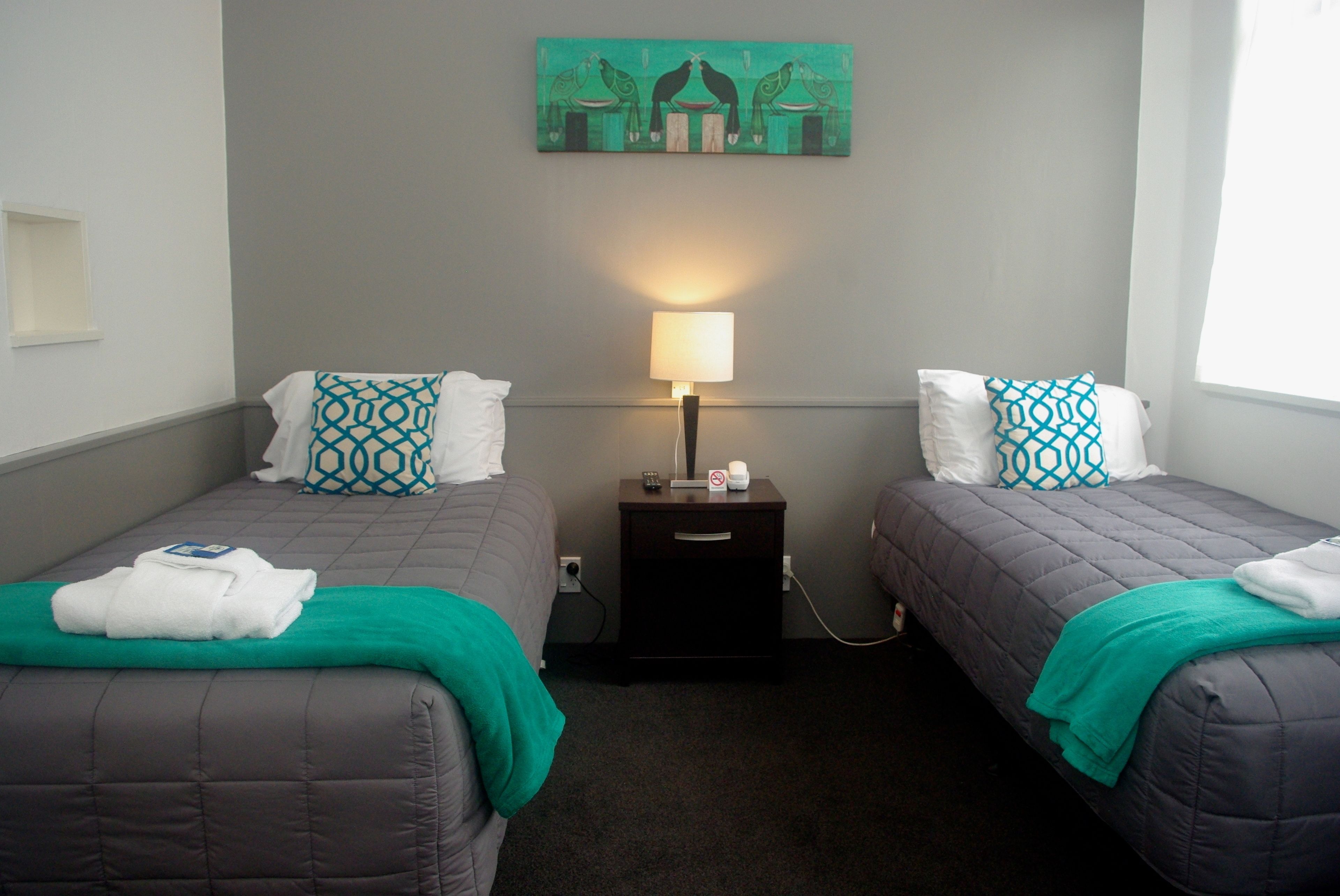 Habitación de hotel con dos camas, cada una con una manta verde y almohadas estampadas, una mesita de noche con lámpara, y un cuadro en la pared.