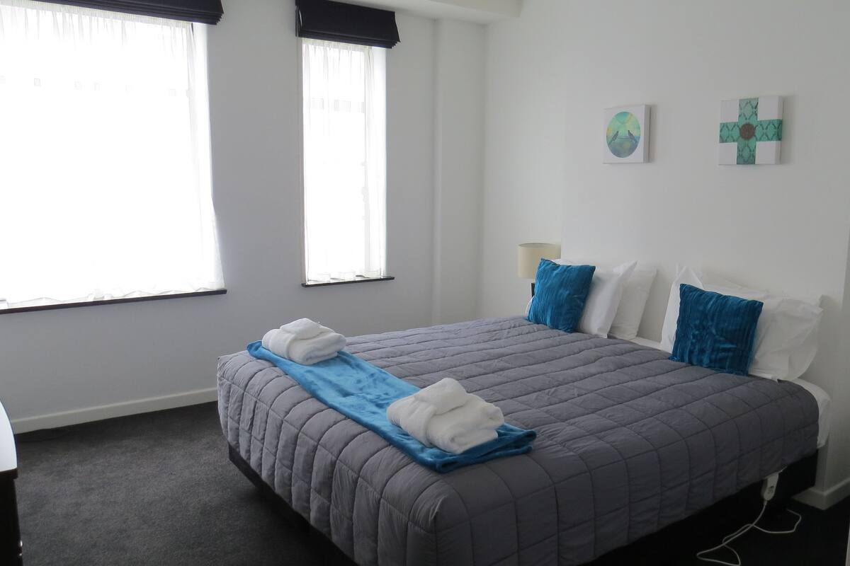A neatly made bed with a blue and white duvet, a grey quilted duvet, and two folded white towels. There are two windows with white curtains and a wall-mounted lamp.