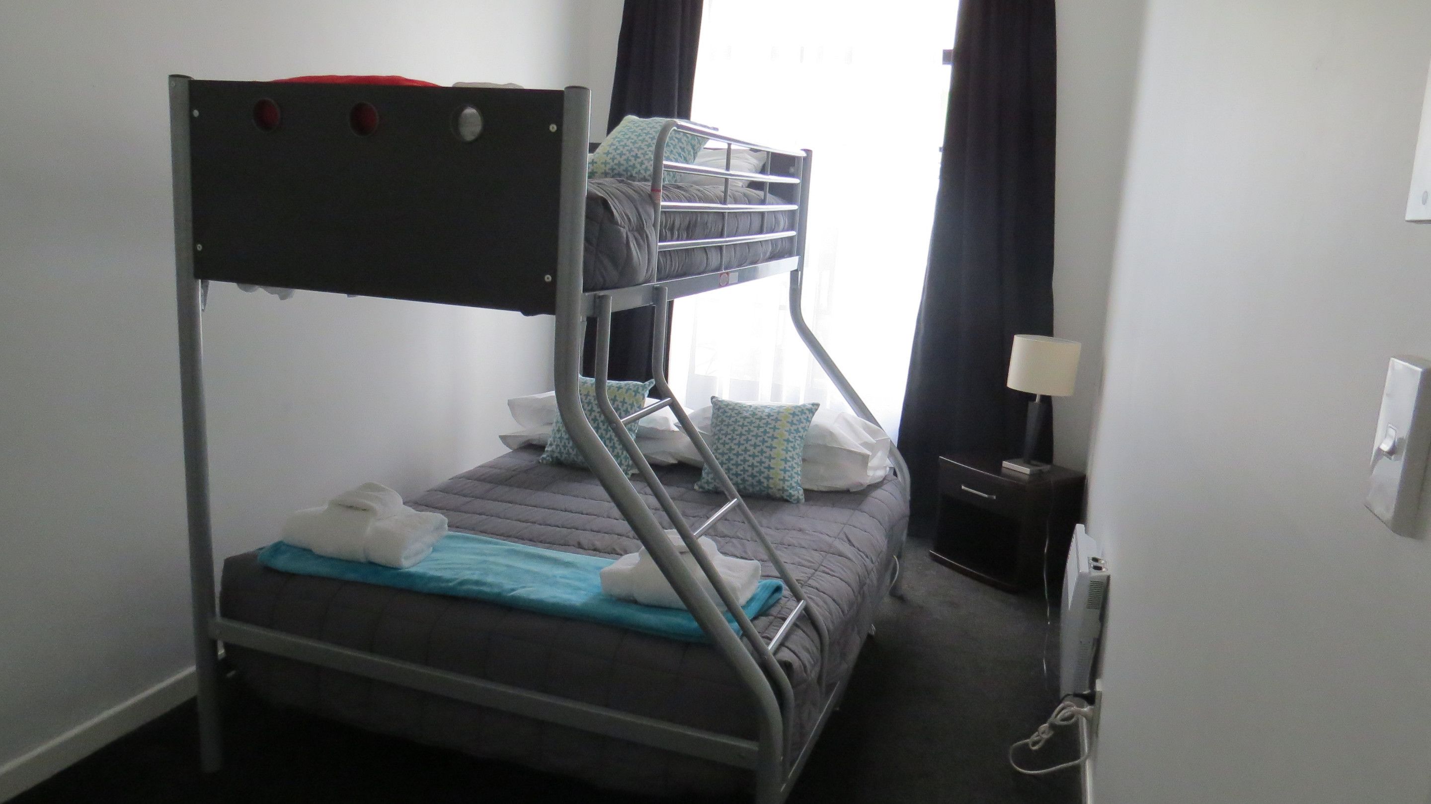 A bunk bed with a metal frame, a pillow, a blanket, and a bedside table with a lamp.