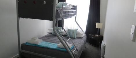 A bunk bed with a metal frame, a pillow, a blanket, and a bedside table with a lamp.