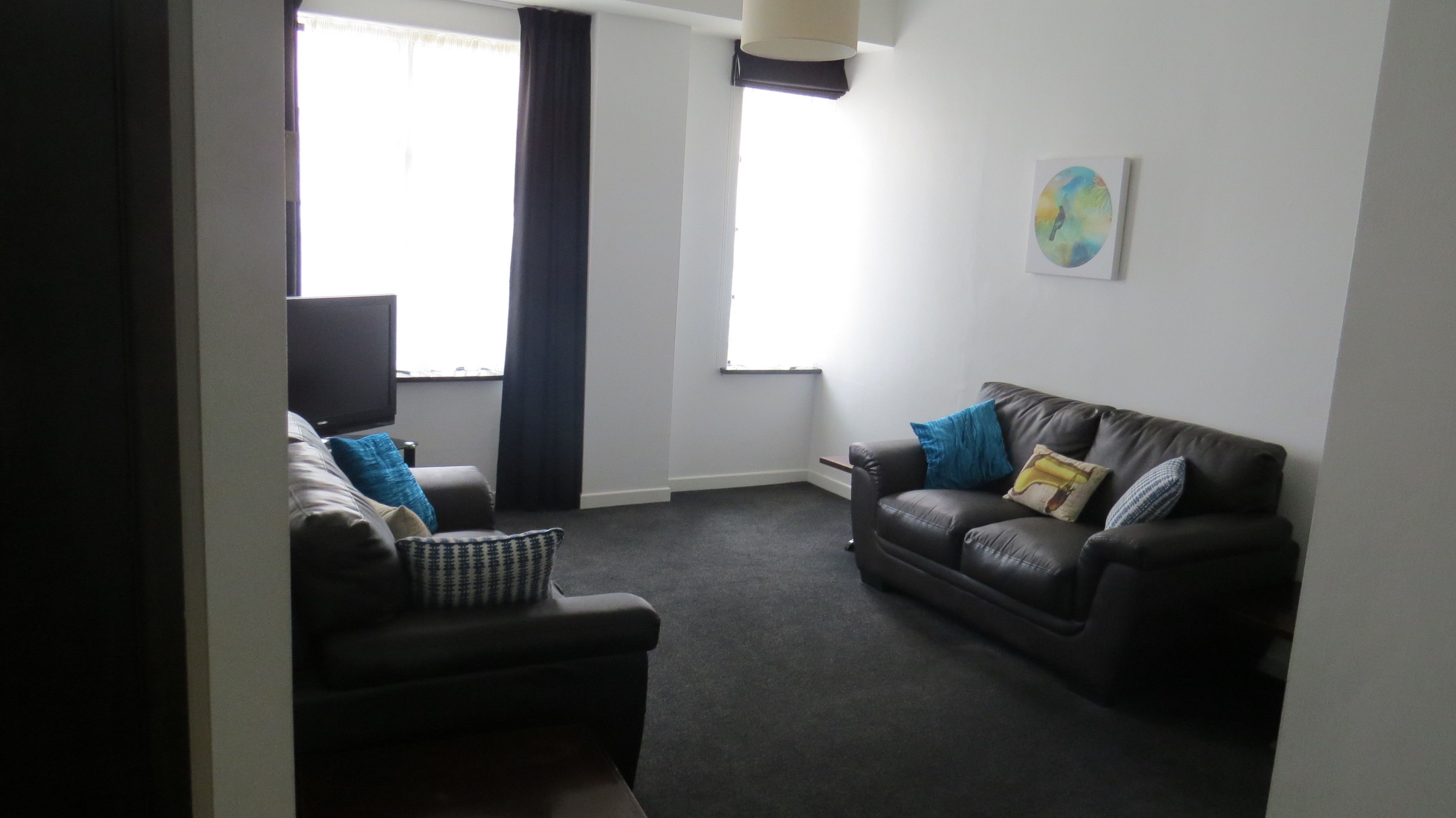 A living room with a dark leather sofa, a small ottoman, and a flat-screen TV. There is a window with curtains and a painting on the wall.