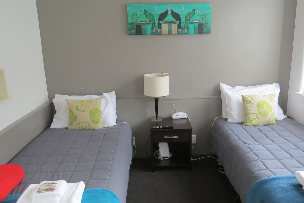 A hotel room with two single beds, a nightstand with a lamp, a telephone, and a painting on the wall.