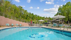 A swimming pool with clear blue water, surrounded by green chairs and umbrellas, and a fence with a view of trees in the background. - Country Town N Suites (Gatlinburg)