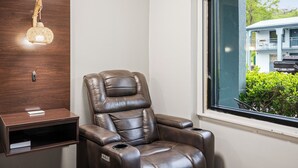 A massage chair in a room with a wooden wall, a small shelf, and a window with a view of a house and greenery. - Country Town N Suites (Gatlinburg)