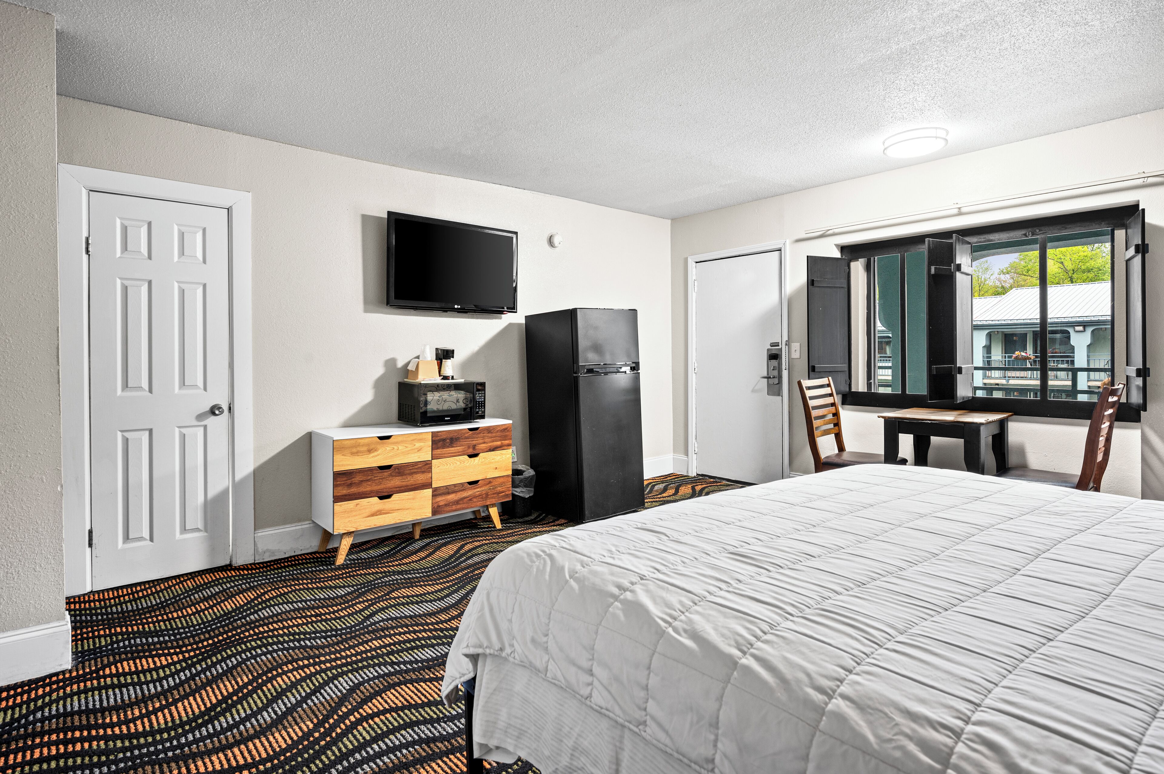 Quarto de hotel com uma cama, televisão na parede, frigorífico, cómoda de madeira e janela com vista para o exterior.