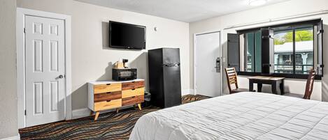 Quarto de hotel com uma cama, televisão na parede, frigorífico, cómoda de madeira e janela com vista para o exterior.