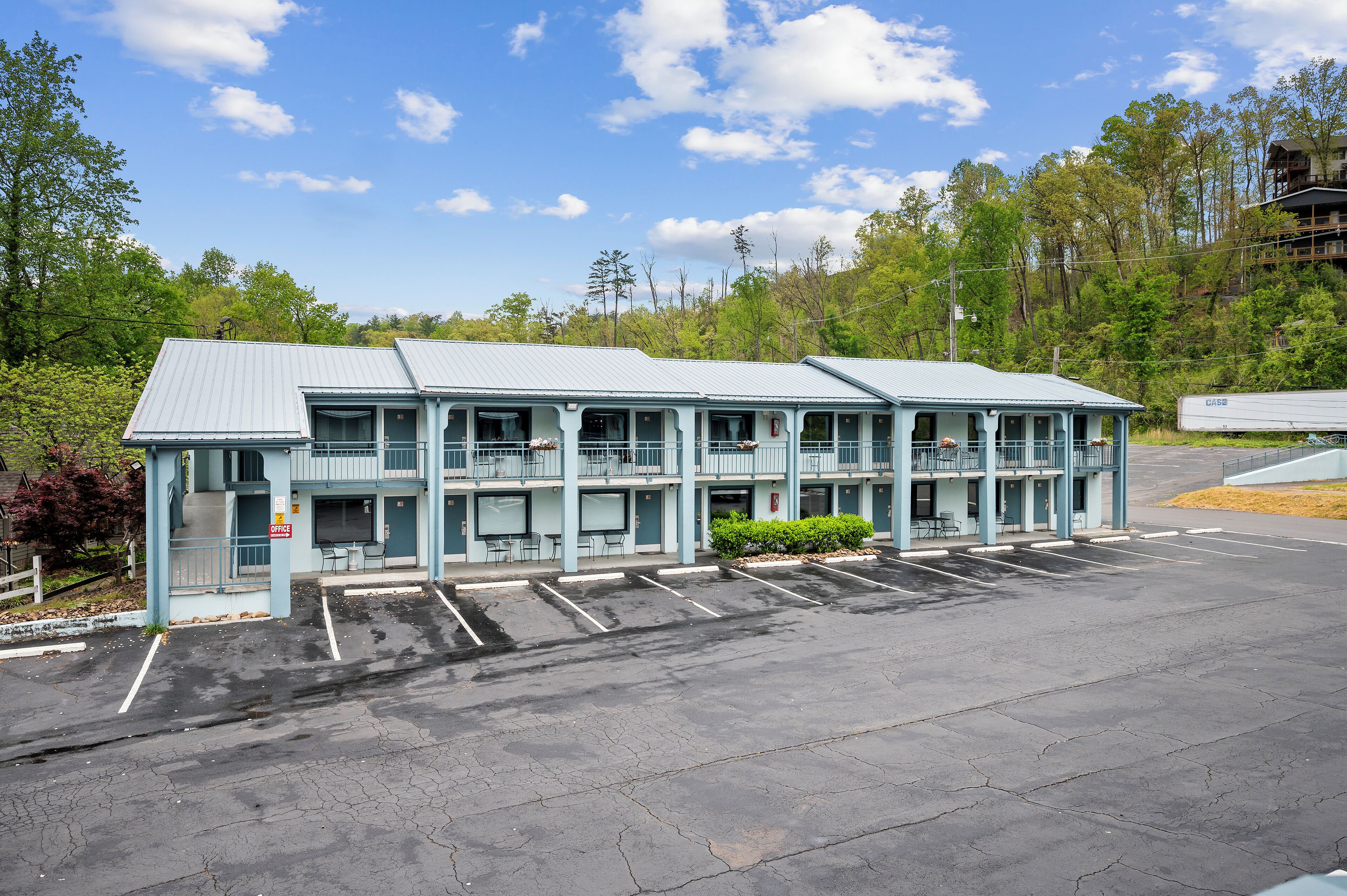 A motel with a row of rooms, a covered parking area, and a view of trees and hills in the background.