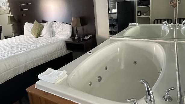 A hotel room with a large bathtub, a bed with white linens, a nightstand with a lamp, and a mirror above the bathtub. - Country Town N Suites (Gatlinburg)