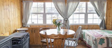L’intérieur d’un chalet accueillant, avec une table à manger en bois, des chaises, un poêle et un lit recouvert d’une courtepointe.