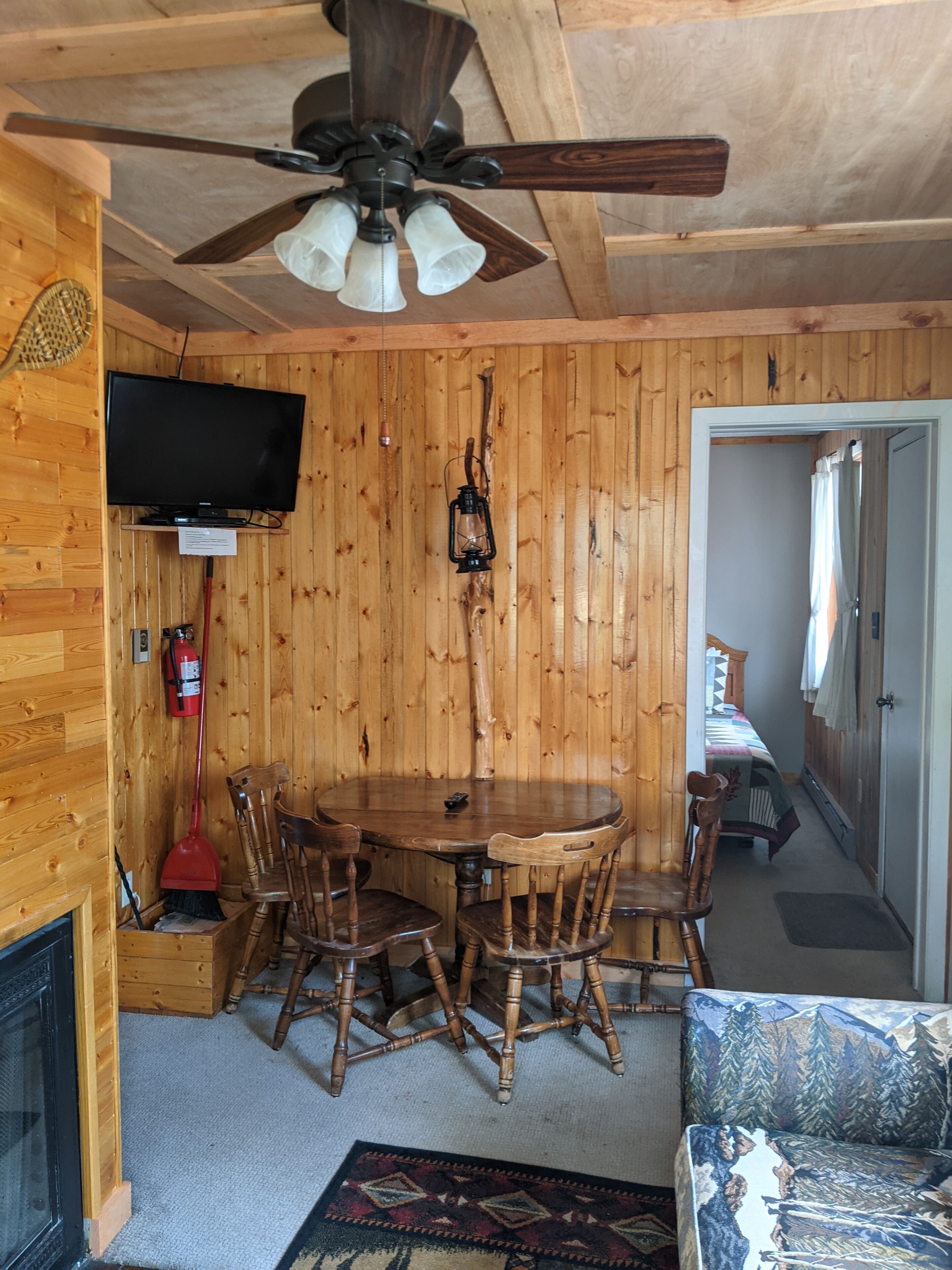 A wooden interior with a round dining table, chairs, a ceiling fan, a mounted TV, and a fire extinguisher.