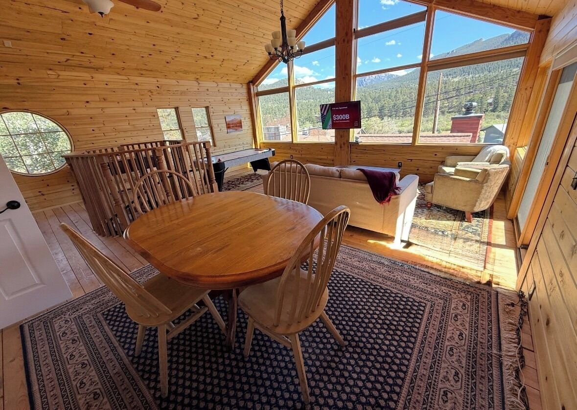 A wooden cabin interior with a dining table, chairs, a sofa, and a view of the outdoors through large windows.