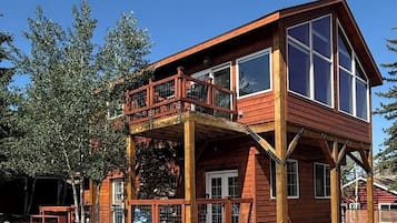A two-story wooden cabin with a balcony, large windows, and a stone foundation.