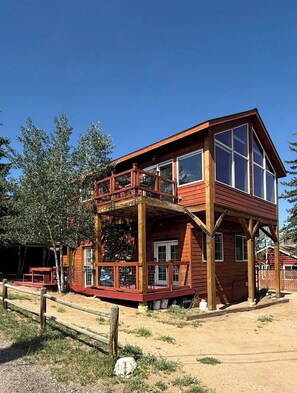 A two-story wooden cabin with a balcony, large windows, and a stone foundation. - Colorado Cottages (Estes Park)
