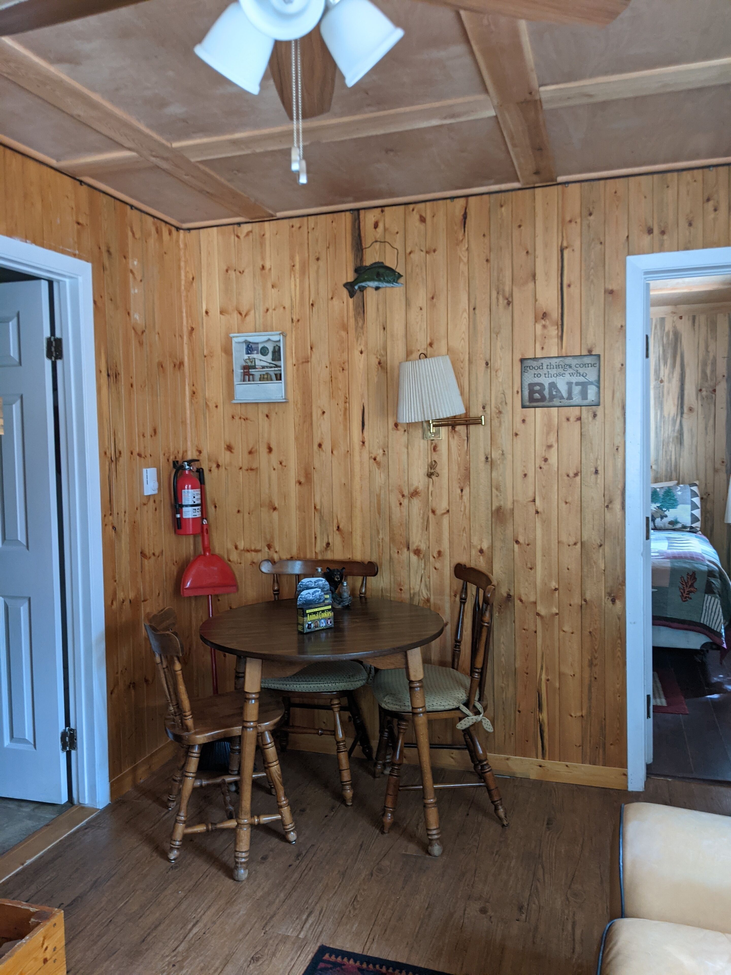 Um quarto com painéis de madeira, uma mesa redonda, cadeiras, um ventilador de teto, um extintor de incêndio e uma cama no quarto vizinho.