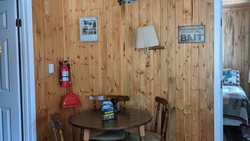 Um quarto com painéis de madeira, uma mesa redonda, cadeiras, um ventilador de teto, um extintor de incêndio e uma cama no quarto vizinho.