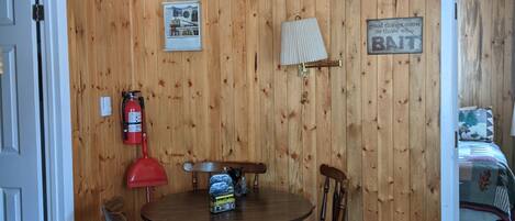 A wooden-paneled room with a round table, chairs, a ceiling fan, a fire extinguisher, and a bed in the adjacent room.
