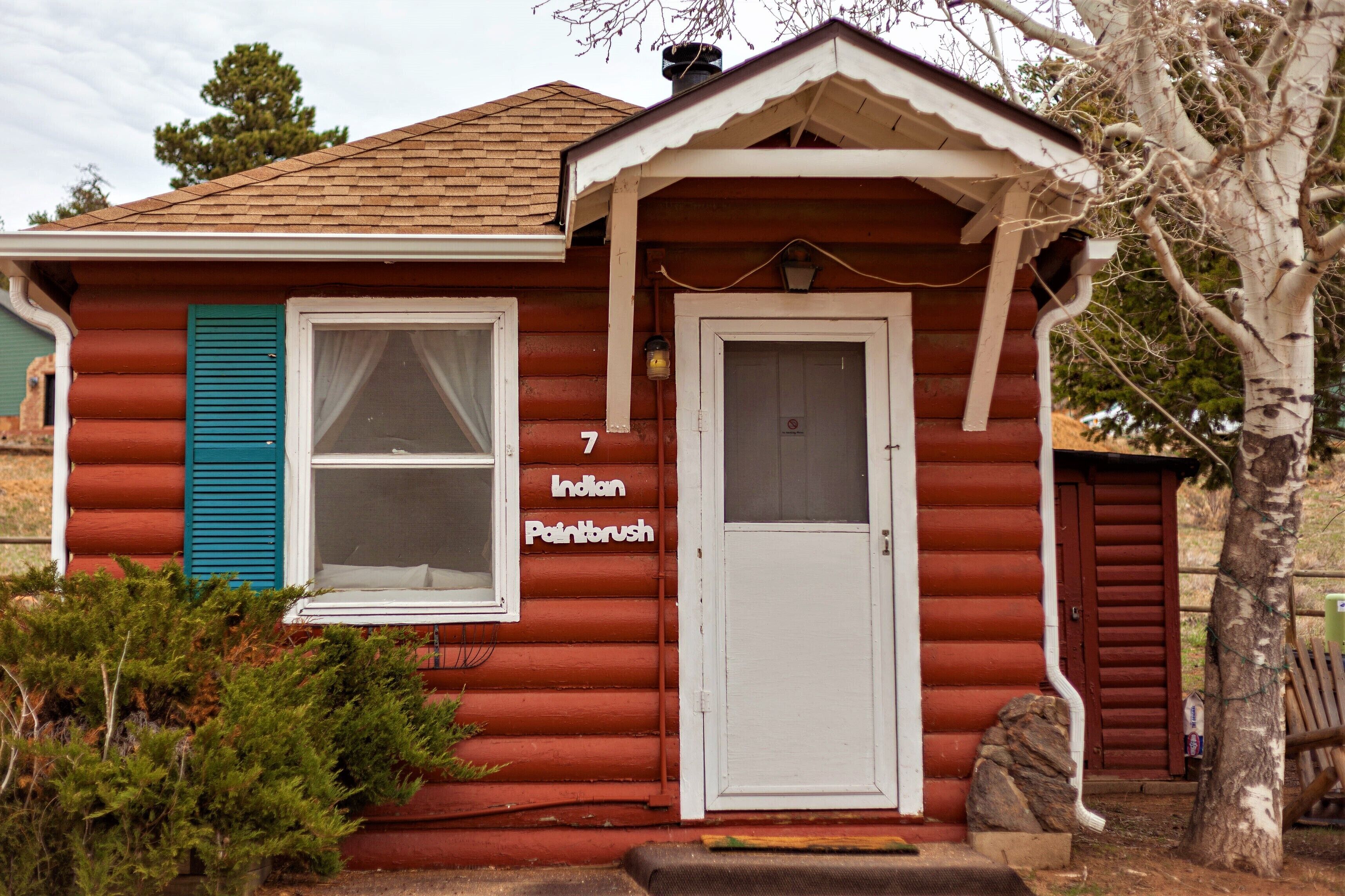 A small, single-story log cabin with a red exterior, a white door, and a window with blue shutters.