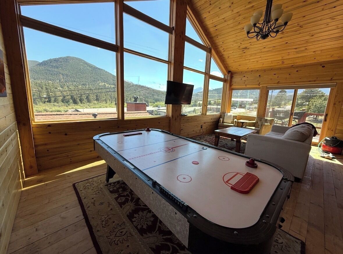 Une pièce au sol en bois, équipée d’une table de billard, d’un canapé et d’un téléviseur. De grandes fenêtres offrent une vue sur un paysage montagneux.