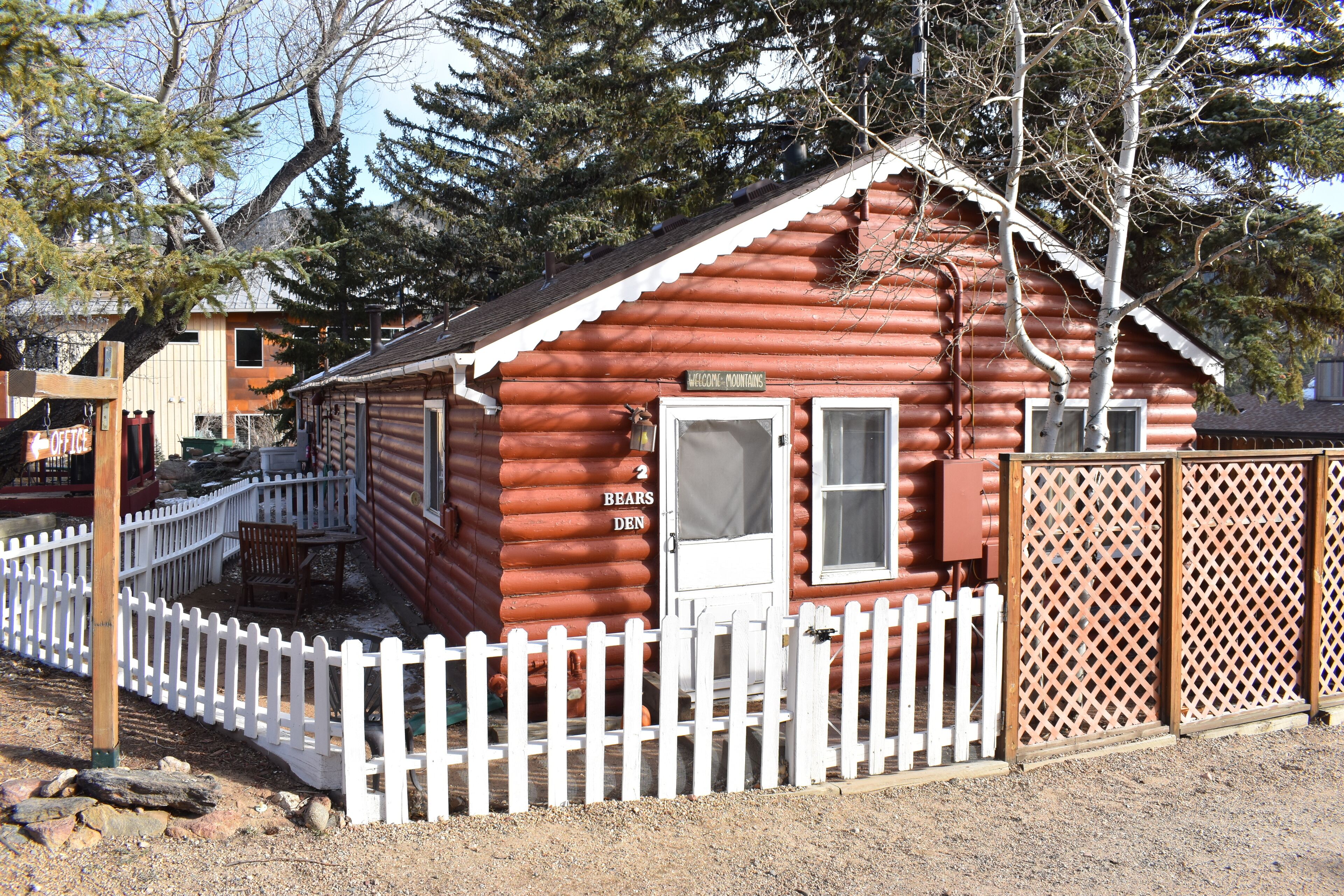 A log cabin with a white picket fence, a door labeled "BEARS DEN," and a sign reading "WELCOME TO FOUNTAIN."