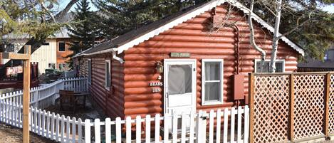 A log cabin with a white picket fence, a door labeled "BEARS DEN," and a sign reading "WELCOME TO FOUNTAIN."