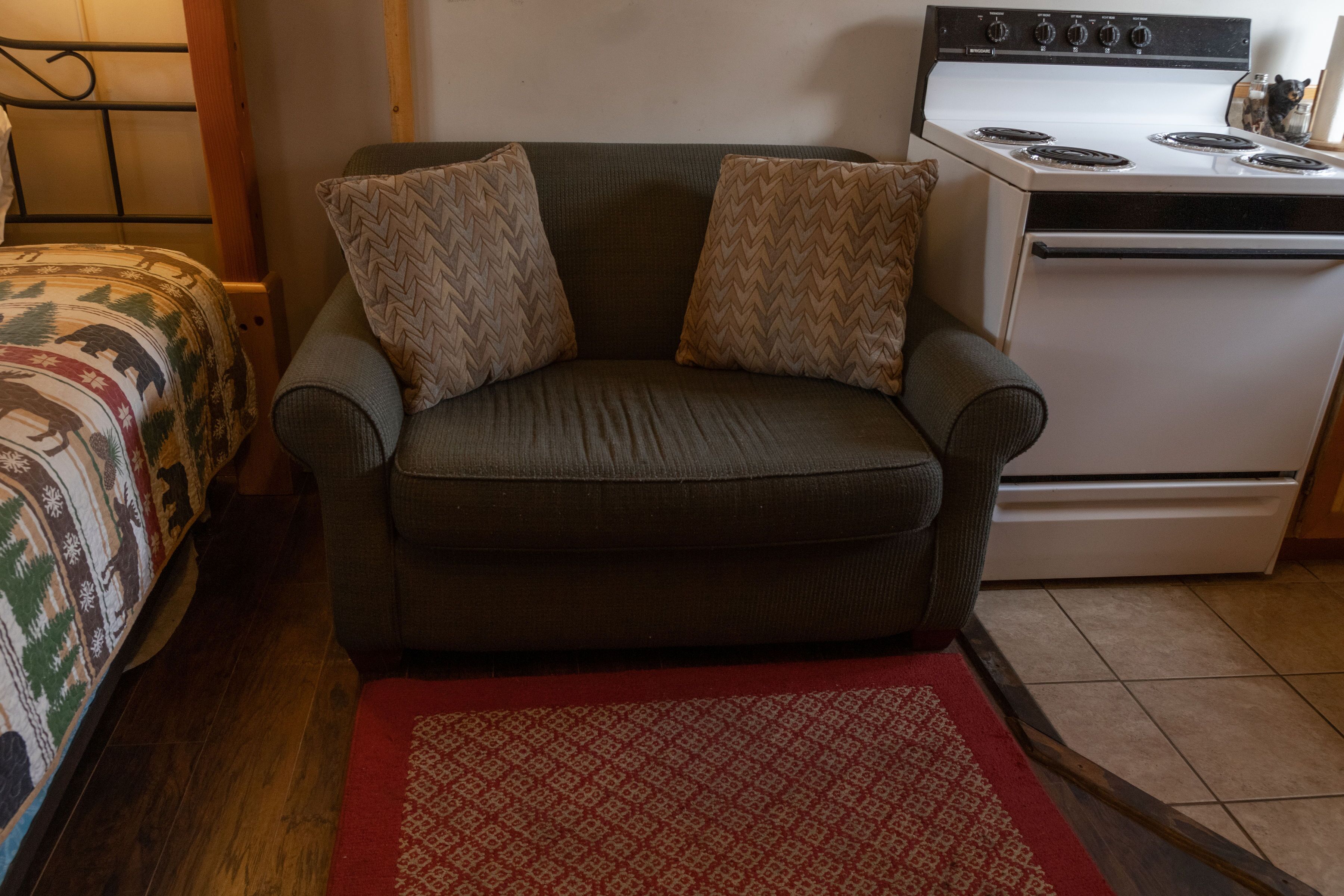 Una habitación pequeña y acogedora con un sofá verde oscuro, dos almohadones estampados, un tapete rojo con diseño, un cabecero de madera y una estufa blanca.