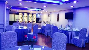 A banquet hall with round tables and chairs, a stage with a backdrop, and a flat-screen TV mounted on the wall. - Summer Palace Hotel (Yangon)