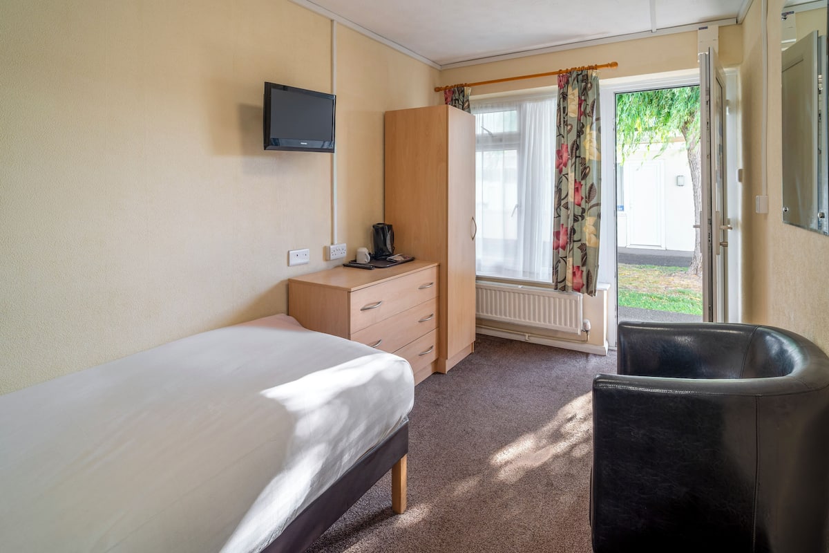 A bedroom with a bed, a wooden wardrobe, a desk with a chair, a television, and a window with curtains.