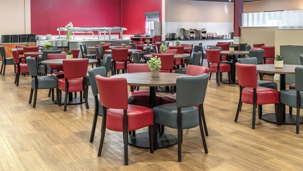 Une cafétéria avec de nombreuses tables et chaises, un espace cuisine à l’arrière-plan et un mur rouge.