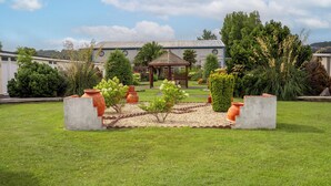 Un jardin bien entretenu, agrémenté d’un belvédère et entouré de diverses plantes et arbres, se trouve devant un bâtiment à la façade blanche et au toit bleu.