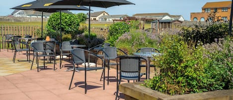 An outdoor patio area with multiple tables and chairs, some under umbrellas, and a variety of plants and flowers.