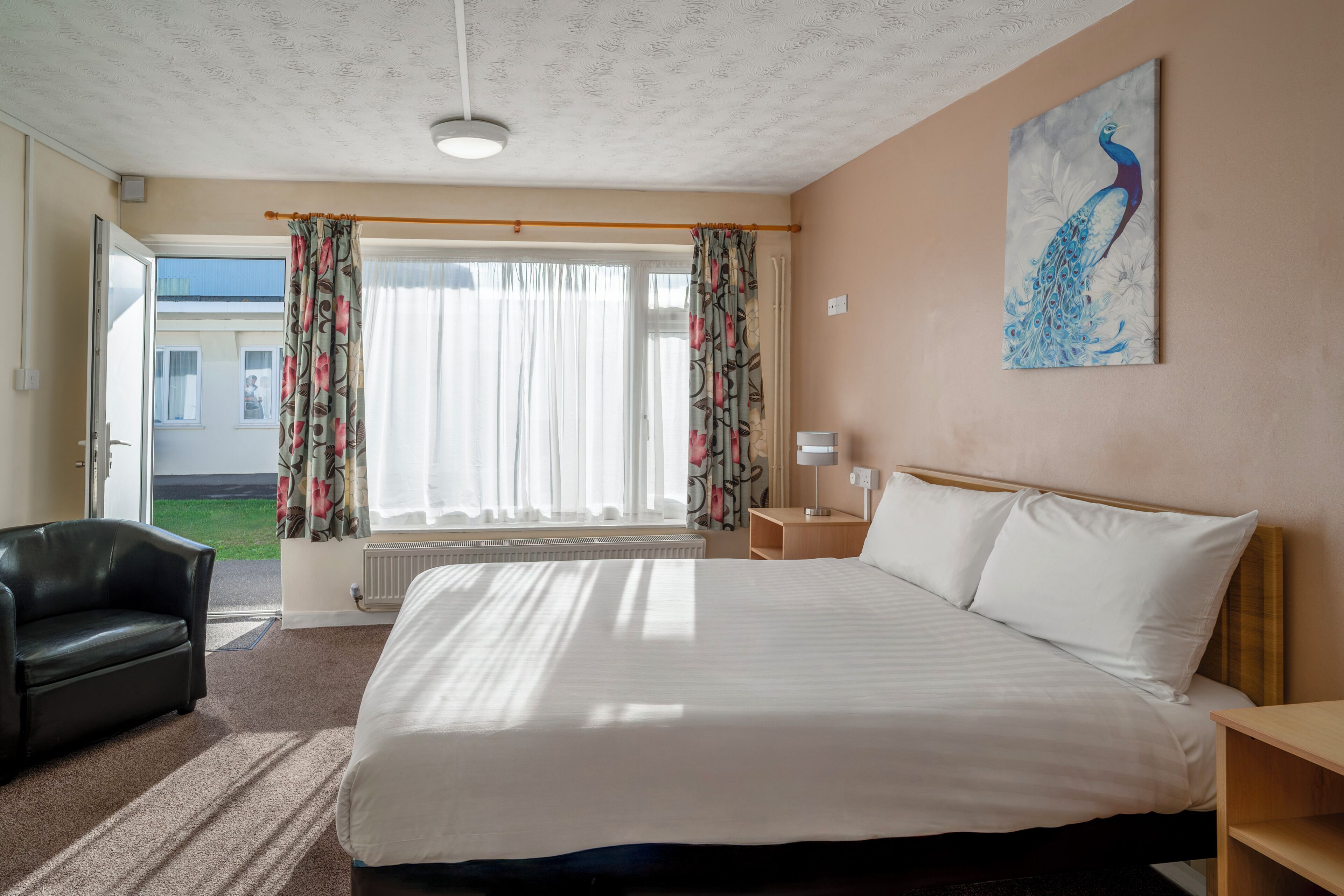 Ein Hotelzimmer mit Bett, Stuhl, Gemälde, einem Fenster mit Vorhängen und Blick ins Freie.