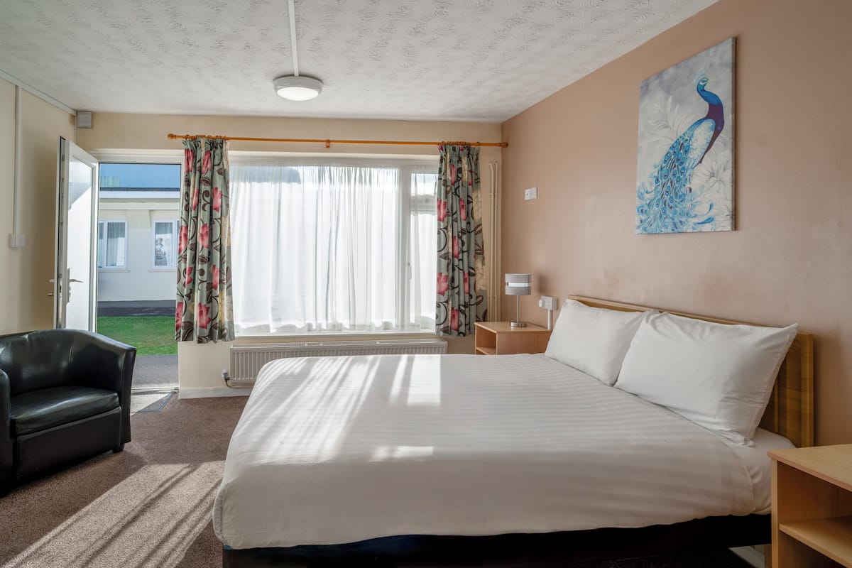 A hotel room with a bed, a chair, a painting, a window with curtains, and a view of the outside.