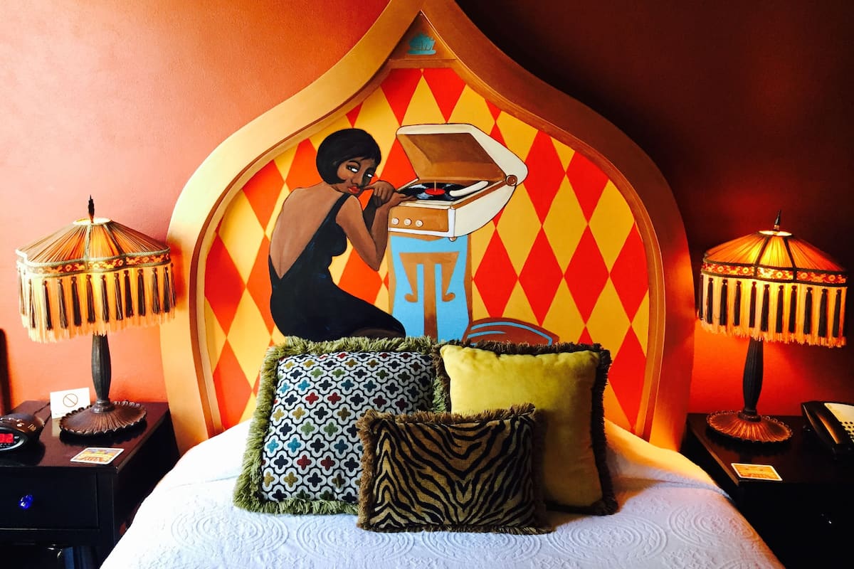 A bed with a headboard featuring a mural of a woman playing a piano. There are three decorative pillows and two lamps on either side of the bed.