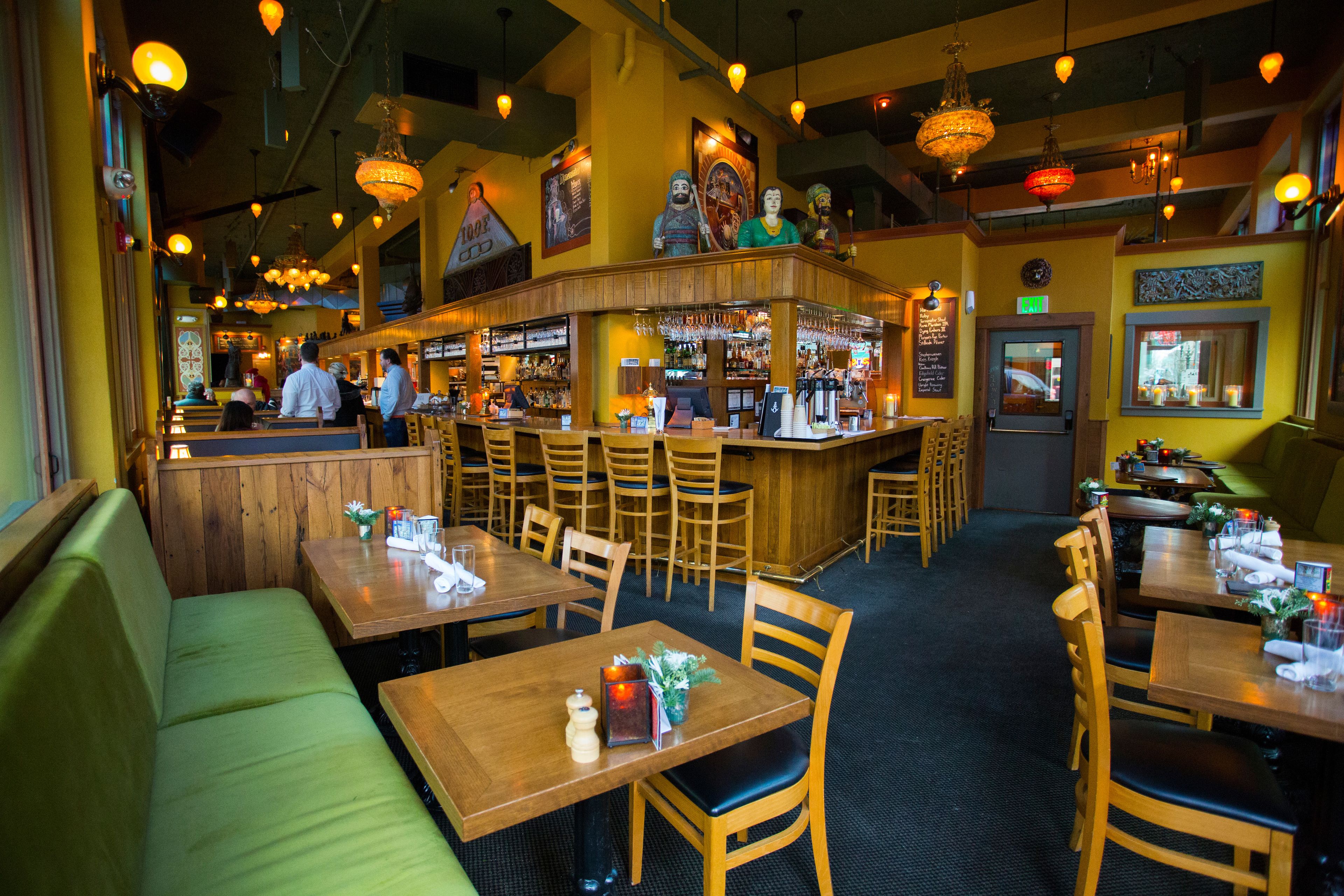 L’intérieur d’un restaurant présente des tables et des chaises en bois, un bar et un espace détente avec des sièges rembourrés de couleur verte.