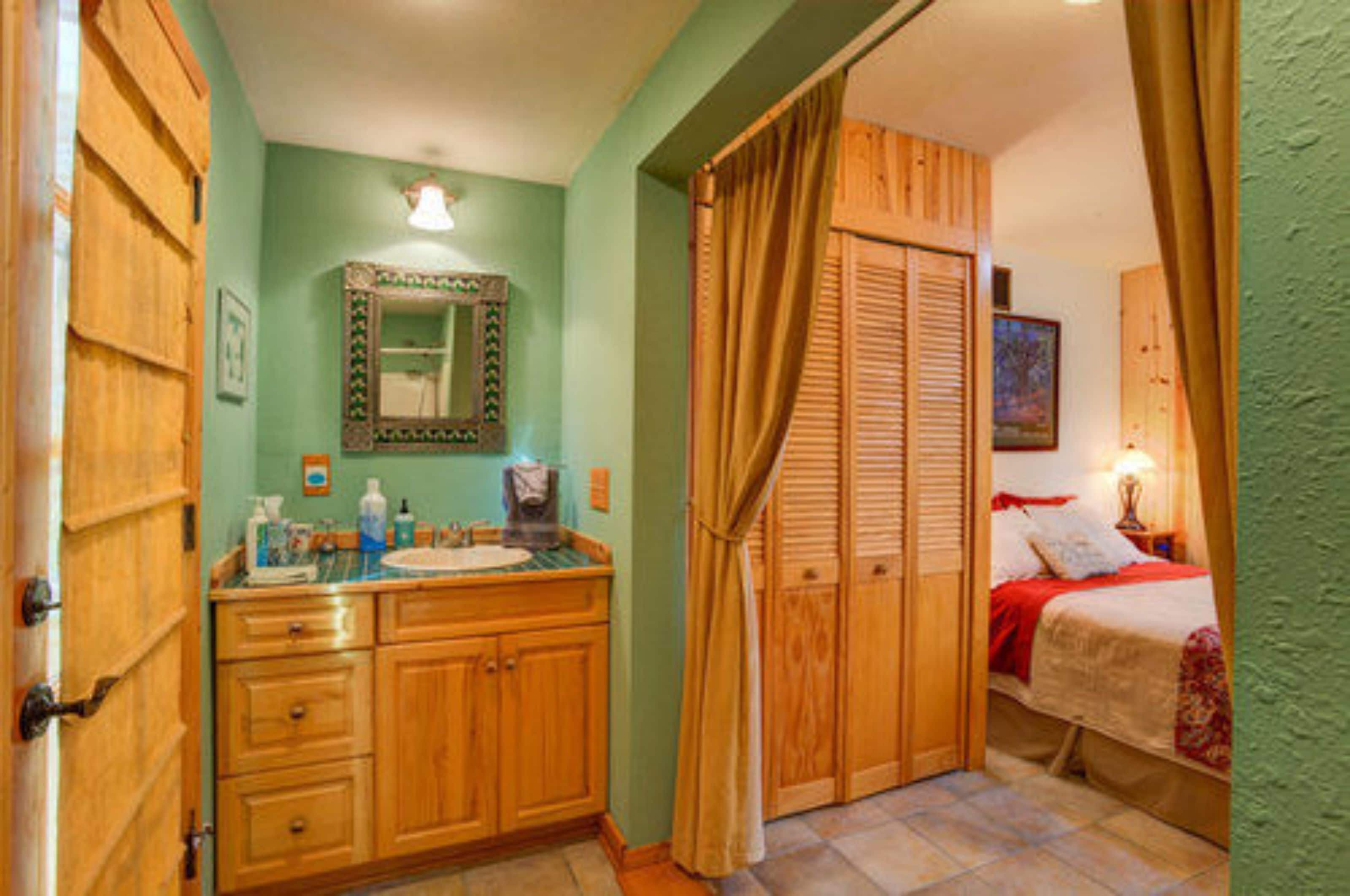 A bedroom with a wooden wardrobe, a bed with red bedding, and a bathroom with wooden cabinets and a mirror.
