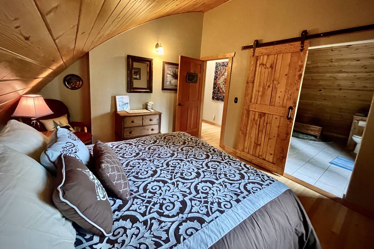 Un dormitorio con techo inclinado, una cama con ropa de cama estampada, un aparador, una lámpara y una puerta que da a un baño.