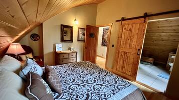 A bedroom with a sloped ceiling, a bed with patterned bedding, a dresser, a lamp, and a door leading to a bathroom.